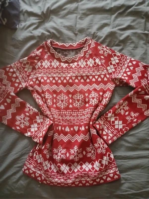 Röd jultröja med snöflingor - Röd stickad tröja med vita snöflingor och klassiskt julmönster. Tröjan har rund halsringning och långa ärmar. Materialet är 98% polyester och 2% elastan vilket gör den mjuk och stretchig. Perfekt för vinterns mysiga dagar.