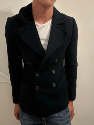 Peacoat - Stilren svart kavaj med dubbelknäppning och breda slag. Klassisk design med fyra knappar framtill och två fickor. Passar perfekt till jeans eller chinos för en snygg look. Materialet ser ut att vara en mjukare tygblandning.