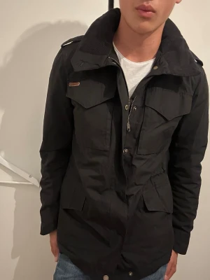 Svart field jacket med hög krage - Stilren svart skepparkavaj med hög krage och axelklaffar. Jackan har flera fickor framtill med lock och dragkedja samt knappar. Materialet ser ut att vara slitstarkt tyg, perfekt för kyligare dagar. Passar dig som gillar en klassisk men modern look.