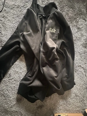 Svart ziptröja från EA7 Emporio Armani - Snygg svart ziptröja från EA7 Emporio Armani med logga på bröstet. Tröjan har hög krage, dragkedja framtill och är tillverkad i ett mjukt material som känns bekvämt. Perfekt för en sportig och stilren look.