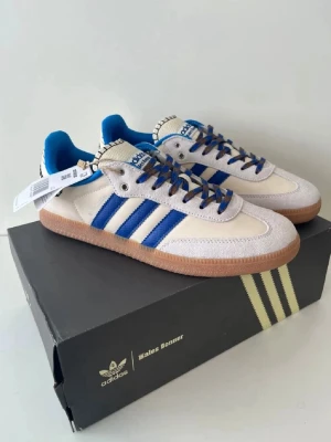 Adidas Wales Bonner skor - Snygga Adidas sneakers i vitt med blå detaljer och klassiska tre ränder på sidan. Skorna har beige mockadetaljer, gummisula och blå/vita skosnören. Insidan är fodrad i blått tyg och hälen har svart/vit grafik. Perfekta för en sportig och trendig look. Aldrig använda, storlek 42.5. Priset är förhandlingsbart vid snabb affär 