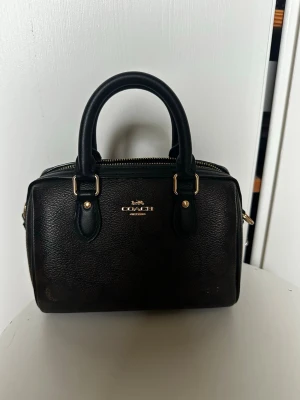 Svart handväska från Coach - Snygg svart handväska från Coach i läder med guldfärgade detaljer och dubbla handtag. Väskan har en klassisk boxig form och dragkedja upptill. Perfekt för dig som gillar stilrena accessoarer med exklusiv känsla.