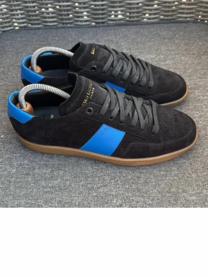  Saint Laurent sneakers  - Snygga svarta sneakers från Saint Laurent i mocka med blåa detaljer på sidan och hälen. Skorna har bruna gummisulor och svarta skosnören. Guldtext på plösen och insidan ger en exklusiv känsla. Perfekta för dig som gillar stilrena och trendiga sneakers.
