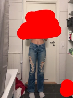 Blå slitna bootcut jeans - Säljer ett par blå bootcut jeans med kraftiga slitningar och hål på framsidan av båda benen. Jeansen har klassisk femficksdesign och normalhög midja. Materialet är denim i bomull och passformen är något utsvängd nedtill.