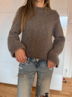 Brun fluffig stickad tröja - Mysig brun stickad tröja med rund hals och långa, lite vidare ärmar. Tröjan har en fluffig struktur och är perfekt för kyligare dagar. Passar till både jeans och kjol för en avslappnad look.