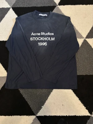 Mörkblå långärmad topp Acne Studios - Mörkblå långärmad topp från Acne Studios med vit text 'Acne Studios STOCKHOLM 1996' på bröstet. Klassisk rund halsringning och avslappnad passform. Perfekt till jeans eller kjol för en clean och stilren look.