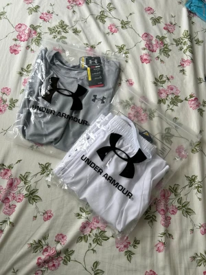 Under Armour träningsset grå/vit - Snyggt träningsset från Under Armour med en ljusgrå t-shirt och vita shorts. T-shirten har stor logga och text på sidan, medan shortsen har resår i midjan, fickor och logga på benet. Perfekt för gymmet eller löpning.