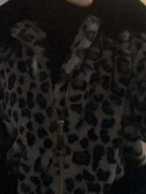 Leopardmönstrad pälsjacka med dragkedja - Säljer en fluffig pälsjacka med leopardmönster i svart och grått. Jackan har en hög krage och stängs med en guldfärgad dragkedja framtill. Perfekt för dig som vill sticka ut och hålla dig varm under vintern.