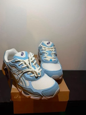 Asics blåvita sneakers - Snygga sneakers från Asics i ljusblått och vitt med klassiska Asics-ränder på sidan. Ovandelen är i mesh och mockaliknande material för en sportig vibe. Skorna har vita snören och en chunky sula i vitt och grått. Perfekta för dig som gillar retrostil och komfort.