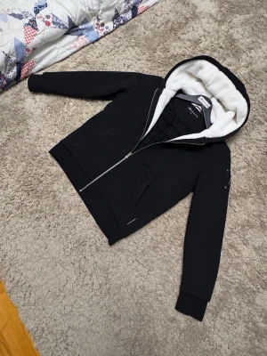 Moose knuckles jacka - Snygg svart hoodie från Mountain Works med dragkedja och stor huva fodrad med vit fuskpäls. Insidan har quiltat mönster och två fickor framtill. Perfekt för dig som vill ha en varm och stilren hoodie med extra mysfaktor.