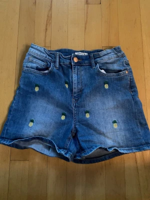 Blå denimshorts med ananasmönster - Snygga blå denimshorts från Lindex med broderade små ananaser över hela plagget. Shortsen har klassisk femficksmodell, knapp och dragkedja framtill samt bälteshällor. Perfekta för sommardagar och ger en lekfull vibe till din outfit.