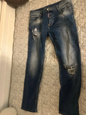 Blå slitna jeans  - Säljer ett par blå jeans från Dsquared2 med slitningar och hål på benen för en riktigt trendig look. Jeansen har fem fickor, knappgylf och klassisk passform. Perfekta för dig som gillar en avslappnad och edgy stil.