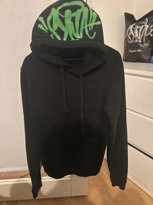Svart syna world hoddie  - Svart hoodie med coolt grön graffiti-tryck på huvan. Klassisk modell med dragsko och ribbade muddar. Perfekt för dig som gillar streetwear och vill sticka ut med en unik detalj på huvan.