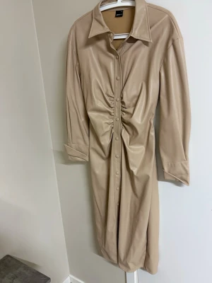Beige långklänning med snörning - Stilren beige långklänning i syntetmaterial med knappar framtill och markerad snörning i midjan. Klänningen har lång ärm, klassisk krage och en rak passform som ger en modern look.