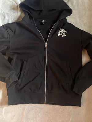 Svart hoodie med tryck och dragkedja - Säljer en svart hoodie från H&M med dragkedja framtill och coolt vitt tryck på bröstet och ryggen. På ryggen står det 'If you don't train to get away then you get caught' runt en tecknad figur. Hoodien har huva och fickor på sidorna.