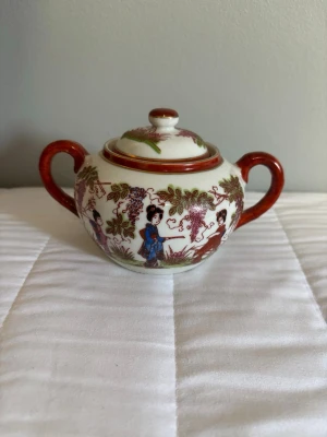 Asiatisk porslinssockerskål med lock - Vacker sockerskål i porslin med asiatiskt motiv, två handtag och lock. Skålen är dekorerad med färgglada figurer, blommor och blad i grönt, rött, blått och brunt. Perfekt som dekoration eller till servering av socker.längd 11cm