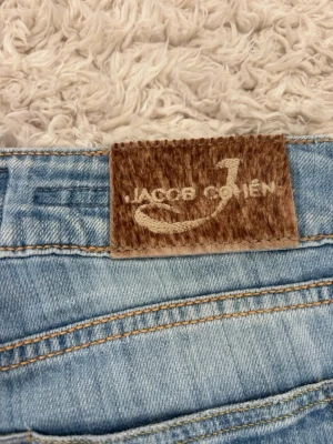 Ljusblå jeans från Jacob Cohen - Säljer ett par ljusblå jeans från Jacob Cohen med klassisk femficksmodell och snygga kontrastsömmar. Jeansen har en patch i mockaimitation med logga bak i midjan och raka ben. Perfekta för en avslappnad och trendig look.