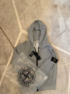 Grå hoodie från Stone Island - Snygg grå hoodie från Stone Island med dragkedja framtill och klassisk logopatch på vänster ärm. Tillverkad i mjuk bomullsmix och har justerbar huva med snören. Perfekt för en avslappnad och trendig stil.