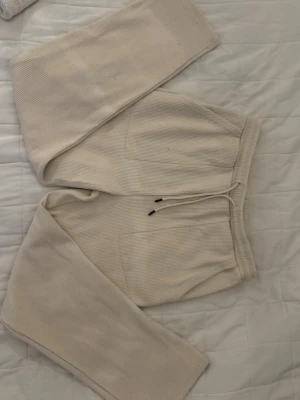 Ribbstickade beige mjukisbyxor - Säljer ett par ribbstickade mjukisbyxor i beige med dragsko i midjan och raka ben. Byxorna har två sidofickor och är gjorda i ett mjukt, stretchigt material som är perfekt för chill dagar hemma.