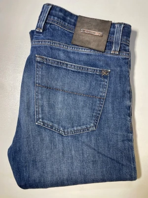 Blå raka jeans från Tramarossa - Snygga blå jeans från Tramarossa med klassisk femficksdesign och raka ben. Jeansen har en diskret logga på bakfickan och patch i läderimitation bak i midjan. Tillverkade i mjukt denimtyg som ger en bekväm passform. Knappen är sönder. Byxorna sitter fortfarande på bra och om du har skärp gör det verkligen ingen skillnad. 