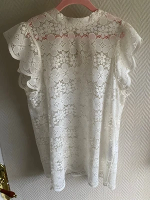 Vit spetsblus med volangärmar - Säljer en superfin vit blus i spets med blommigt mönster och volangdetaljer på ärmarna. Blusen är ärmlös med rund halsringning och knapp i nacken.   Har även den i svart så man kan köpa båda för300