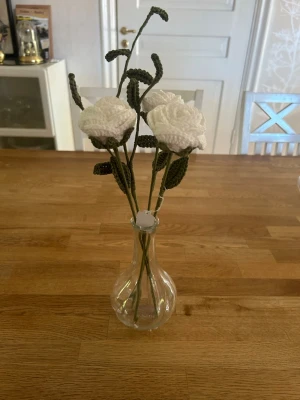 Virkade blommor i vas - Unika handgjorda virkade rosor i vitt garn med gröna stjälkar och blad. Blommorna står i en enkel, genomskinlig glasvas. Perfekt som dekoration till hemmet eller som present till någon speciell.
