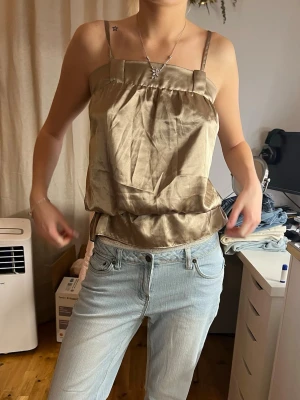 Champagnefärgad linnetopp i satin - Snygg champagnefärgad linnetopp i glansigt satinmaterial. Toppen har tunna axelband och en avslappnad passform med lätt puffig effekt nedtill. Perfekt att styla med jeans eller kjol för en trendig look.