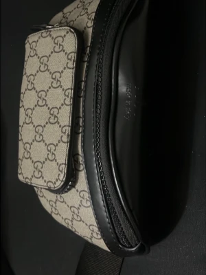 Beige shoppingväska från Guc Ci  - Säljer en beige shoppingväska från Guc Ci med det klassiska guc Ci mönstret över hela väskan. Väskan har svarta detaljer i läder och dragkedja. Perfekt för dig som vill ha en stilren och lyxig accessoar till din outfit.