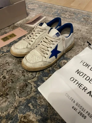 Golden Goose vita sneakers med blå stjärna - Golden Goose sneakers i vitt läder med blå detaljer och ikonisk blå stjärna på sidan. Låga med perforerad tå och beige sula. Skorna har vita snören och blå insida, perfekt för dig som gillar en unik och trendig look.