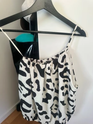 Svartvit one shoulder topp med djurmönster - Säljer en luftig svartvit topp one shoulder med coolt djurmönster och tunna axelband. Toppen har resår både upptill och nertill för en avslappnad passform. Perfekt för varma dagar och enkel att styla med jeans eller kjol.