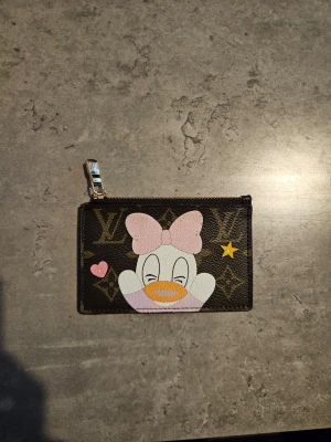 Louis Vuitton plånbok med Daisy Duck - Snygg brun plånbok från Louis Vuitton med klassiskt monogrammönster och ett tryck av Daisy Duck med rosa rosett. Plånboken har guldfärgad dragkedja och detaljer i läder. Perfekt för kort och mynt.