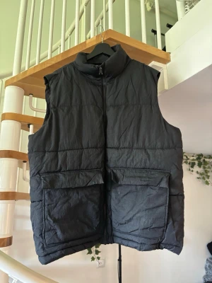 Svart dunväst med stora fickor - Säljer en svart dunväst med hög krage och dragkedja framtill. Västen har två stora fickor framtill och quiltad design som ger en puffig look. Perfekt för lager-på-lager och riktigt skön att bära under kyliga dagar. Västen har ett väldigt litet brännmärke på vänster sida