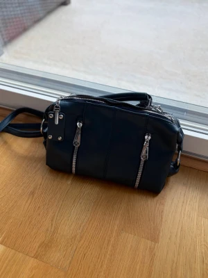 Mörkblå  axelväska med silverdetaljer - Snygg mörkblå axelväska i slätt material med flera silverfärgade dragkedjor och detaljer. Väskan har justerbar axelrem och praktiska fack framtill. Perfekt för dig som gillar en stilren och modern look.                     ( handtaget har låstnat men går att sätta tillbaka)
