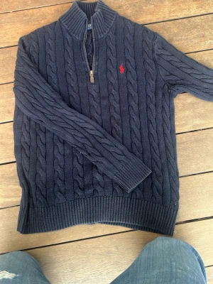 Mörkblå kabelstickad tröja Polo - Mörkblå kabelstickad tröja från Polo Ralph Lauren med halv dragkedja och hög krage. Tröjan har ett rött broderat logomärke på bröstet och ribbade muddar vid ärmslut och nederkant. Perfekt för lager på lager och klassisk stil.