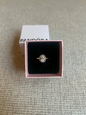 Guldfärgad ring med stenar från Pandora - Snygg guldfärgad ring från Pandora med en stor, glittrande oval sten i mitten och små klara stenar runt om. Ringen har en klassisk och elegant design som verkligen sticker ut. Perfekt accessoar för att lyfta din stil.