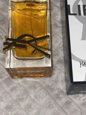 Yves Saint Laurent Libre parfym - Säljer en elegant parfym från Yves Saint Laurent, Libre. Flaskan är rektangulär i tjockt, genomskinligt glas med guldiga detaljer och logotyp framtill. Innehållet är bärnstensfärgat och flaskan har en lyxig och modern design.