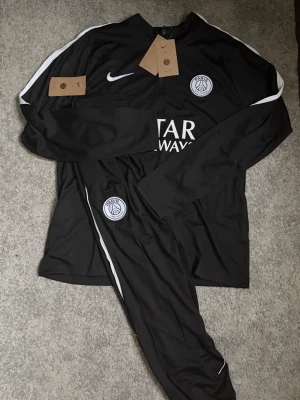 PSGTracksuit - Säljer ett svart träningsset med långärmad tröja och matchande byxor. Setet har vita detaljer längs ärmar och ben,  på bröstet och byxan. Materialet är lätt och andas, perfekt för fotbollsträning.