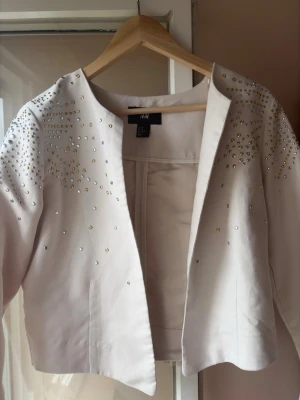 Ljusbeige jacka med fransar och nitar - Snygg ljusbeige jacka från H&M med långa ärmar, dekorerad med silvriga och guldiga nitar på axlar och överdel. Baksidan har långa fransar som ger en cool och trendig look. Jackan är öppen framtill och har en kort, boxig passform.