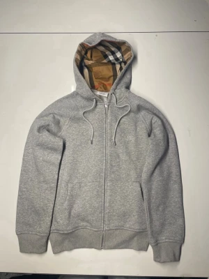 Grå hoodie med dragkedja och mönstrad huva - Snygg grå hoodie med dragkedja och fickor framtill. Huvan har ett klassiskt rutigt mönster på insidan och justerbara snören. Tillverkad i mjukt sweatshirtmaterial, perfekt för en avslappnad stil.
