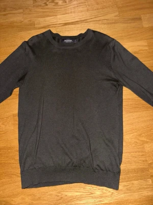 Mörkgrön stickad tröja Dressmann - En stilren mörkgrön stickad tröja från Dressmann i storlek S. Tröjan har rund halsringning, långa ärmar och ribbade muddar vid ärmslut och nederkant. Tillverkad i mjuk bomull för en bekväm känsla.