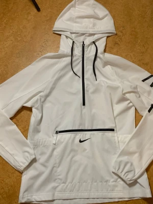 Vit vindjacka med huva från Nike - Snygg vit vindjacka från Nike med svart dragkedja och logga på magen. Jackan har huva med dragsko, stor ficka framtill och elastiska muddar vid ärmsluten. Perfekt för blåsiga dagar och sportiga outfits.