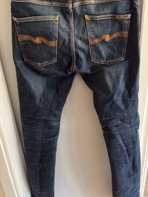 Mörkblå smala jeans Nudie Jeans - Säljer ett par mörkblå smala jeans från Nudie Jeans med klassisk femficksmodell och kontrastsömmar. Jeansen har en tight passform och är tillverkade i denim med snygg slitning på låren och knäna. Perfekta för en avslappnad och trendig look.