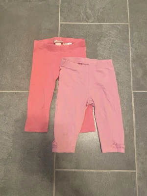 Två par rosa trekvarts leggings med rosett - Säljer två par trekvarts leggings i olika nyanser av rosa. Ena paret är ljusrosa med en söt liten rosett vid benslutet, det andra paret är mer mörkrosa. Båda har resår i midjan och är tillverkade i mjukt, stretchigt bomullstyg. Perfekta för en bekväm och avslappnad stil.