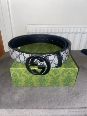 Gucci bälte med GG-spänne - Snyggt Gucci bälte med klassiskt GG-mönster i vitt och blått. Bältet har ett stort, rundat GG-spänne i svart metall och tydlig logga längs bandet. Perfekt accessoar för att lyfta vilken outfit som helst.