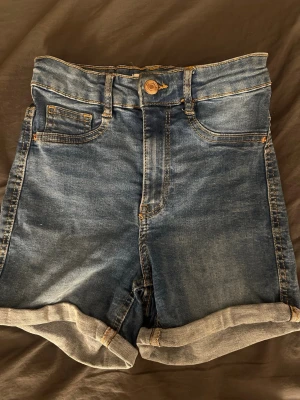 Blå högmidjade jeansshorts - Säljer ett par blå högmidjade jeansshorts från Molly med klassisk femficksdesign och uppvikta benslut. Shortsen har knapp och dragkedja framtill samt bälteshällor. Perfekta till sommaren och lätta att matcha med olika toppar.