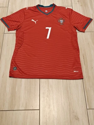 Portugal hemmaställ 2026  - Snygg röd Portugal fotbollströja från Puma med nummer 7 och Ronaldo på ryggen. Tröjan har gröna och vita detaljer vid krage och ärmslut samt vågigt mönster i tyget. Tillverkad i lätt funktionsmaterial med DryCell-teknologi.