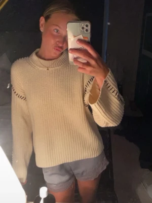 Beige stickad tröja med detaljer - Säljer en beige stickad tröja med bred ribbstickning och coola håldetaljer längs ärmarna. Tröjan har en högre rund halsringning och långa ärmar med avslappnad passform. Perfekt för dig som gillar en enkel men ändå unik look.
