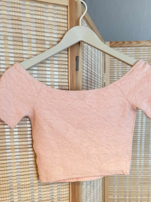 Ljusrosa/persikofärgad croppad topp med struktur - Söt ljusrosa croppad topp med korta ärmar och offshoulder-design. Toppen har en strukturerad yta som ger en unik look och är perfekt för dig som gillar trendiga detaljer. Passar till både jeans och kjol för en cool stil.