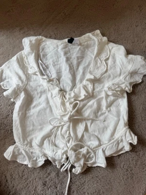 Vit blus med snörning och volanger - Supersöt vit blus i linne med korta ärmar, volangkant och snörning framtill. Blusen har v-ringning och volanger längs halsringningen och nederkanten för en romantisk vibe. Perfekt till jeans eller kjol för en trendig look. Just nu ser del väldigt skrynklig ut men det är bara att stryka🥰