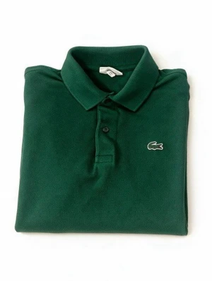 Grön Lacoste pikétröja  - Mörkgrön pikétröja från Lacoste med klassisk krage och två knappar framtill. Tröjan har det ikoniska krokodilmärket broderat på bröstet och är tillverkad i bomull. Perfekt för en clean och stilren look.